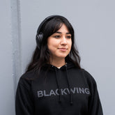 Person wearing black headphones and a black hoodie with 'BLACKWING' logo against a gray wall.