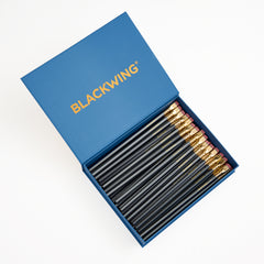 A set of Blackwing 602 wooden graphite pencils with gold ferrules and pink erasers, neatly arranged inside an open blue box with the brand name BLACKWING printed in gold on the lid, shot from a top-down angle on a white background.