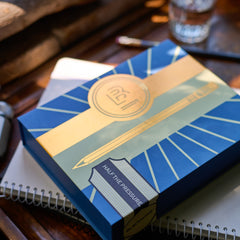 A square box with a geometric art-deco style cover design in blue, yellow, and beige, featuring a pencil illustration and a black spine band labeled 'HALF THE PRESSURE, BLACKWING.'.