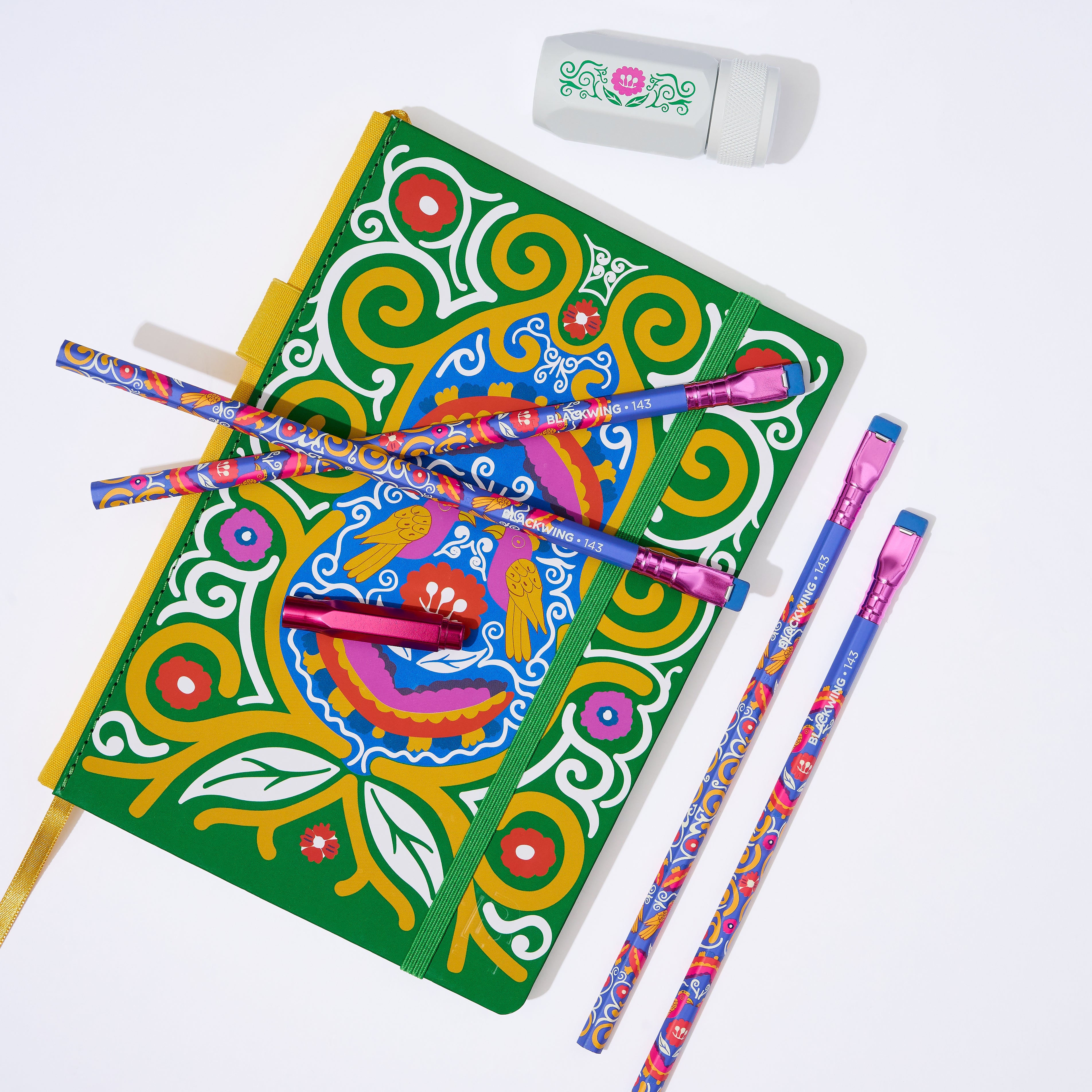 A colorful decorative notebook with a green cover and ornate multicolor swirl and floral patterns, shown with four matching patterned pencils, a pink metallic point guard, and a white eraser on a clean white background.