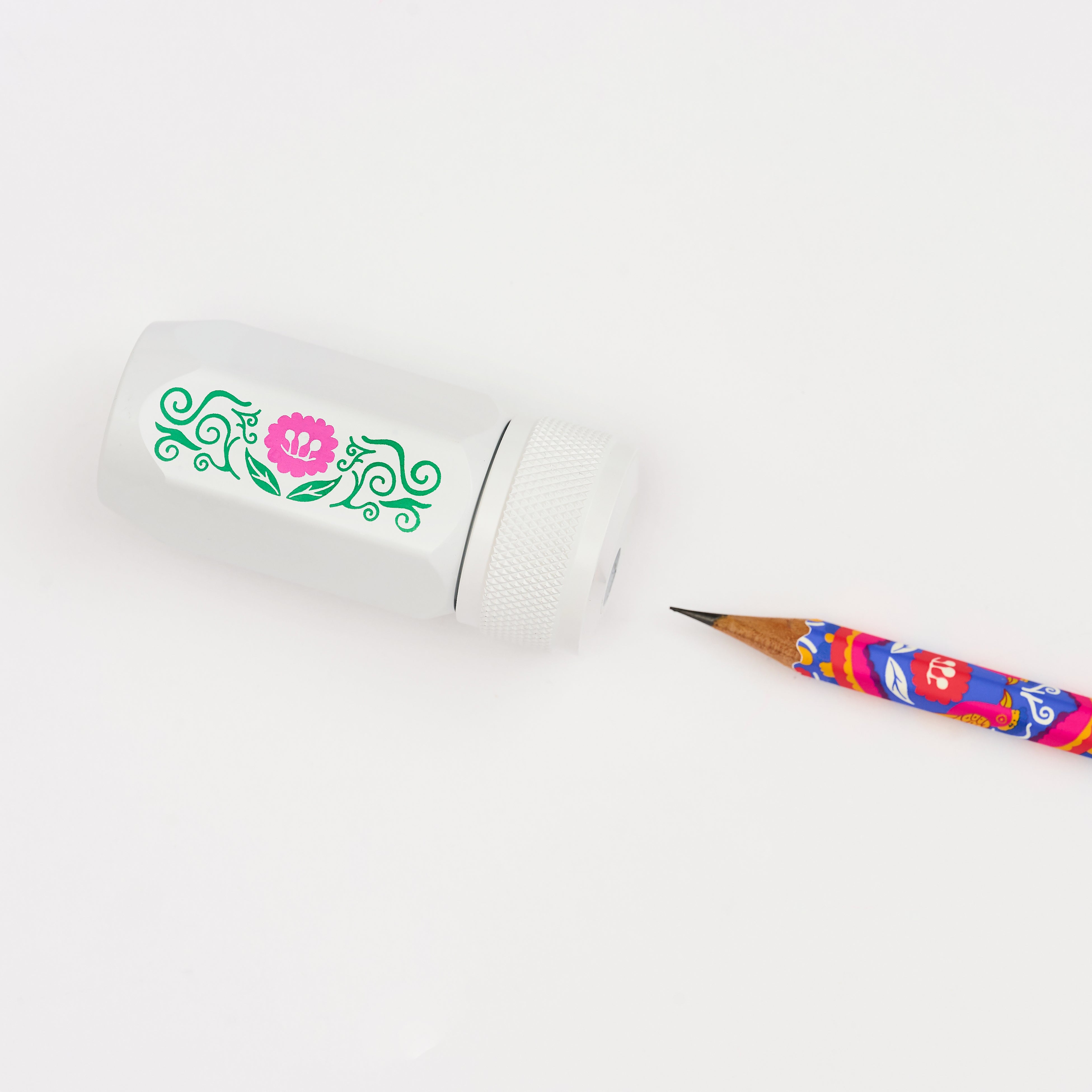 A white pencil sharpener with a green and pink floral design, shown next to a colorful pencil on a clean white background.