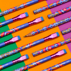 A set of colorful wood pencils arranged diagonally on a vibrant, colorblock background of orange, green, and pink blocks. The pencils feature blue bodies with a busy, multicolored floral and swirl pattern, metallic pink erasers, and white text reading 'BLACKWING 143', in honor of Frida Kahlo
