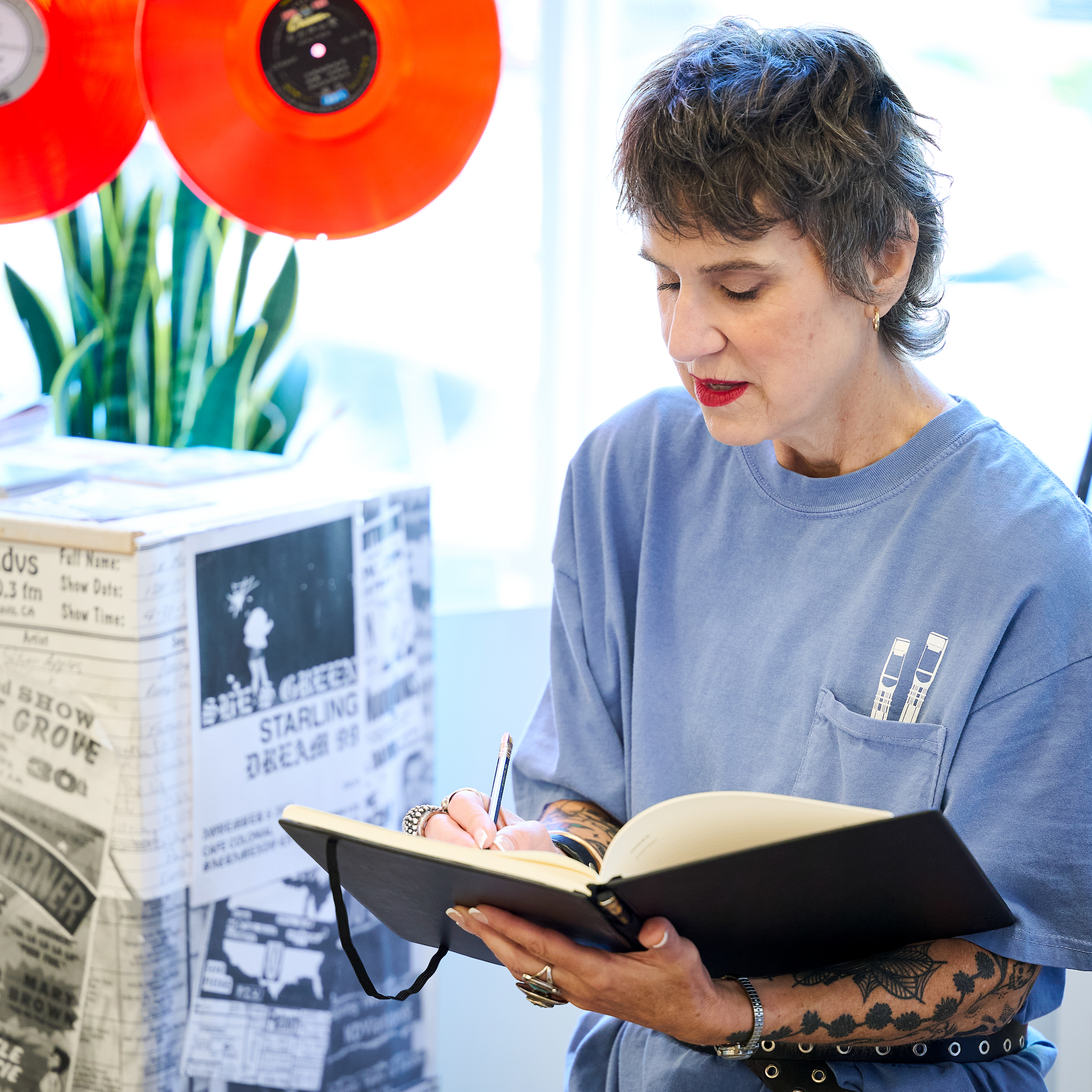 Woman wearing a blue t-shirt writing in a hardcover notebook with a pencil.