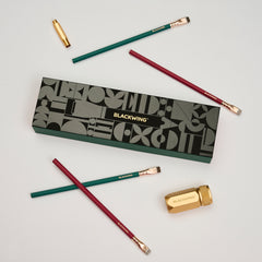 Blackwing Holiday Essentials Gift Set Box surrounded by the contents, including 2 green pencils, 2 red pencils, a gold sharpener, and a gold point guard on a white background.