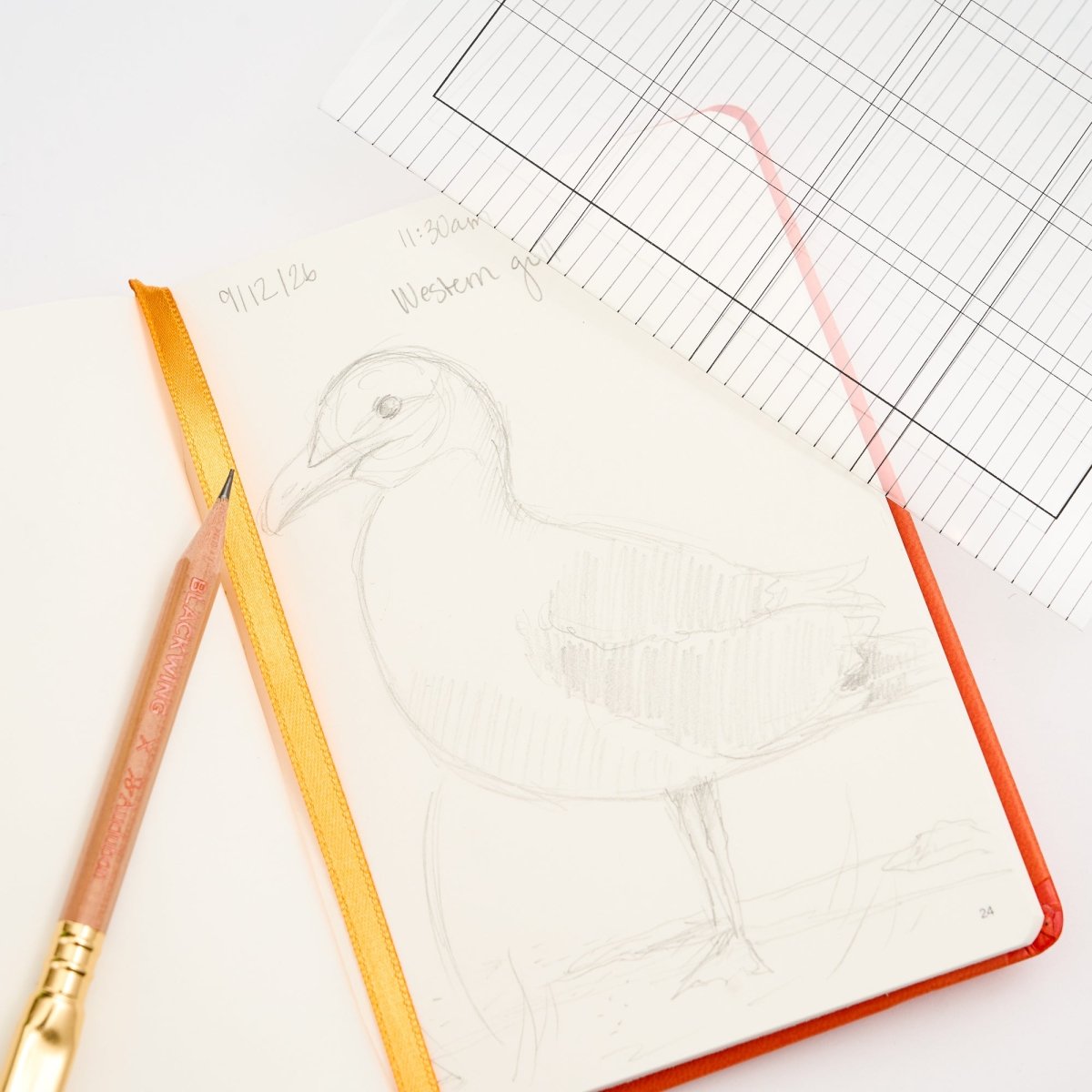 An open sketchbook with a pencil drawing of a bird, featuring a bright orange cover and a yellow-orange ribbon bookmark. A Blackwing pencil and lined paper sheets are arranged on a white background.
