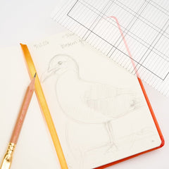 An open sketchbook with a pencil drawing of a bird, featuring a bright orange cover and a yellow-orange ribbon bookmark. A Blackwing pencil and lined paper sheets are arranged on a white background.