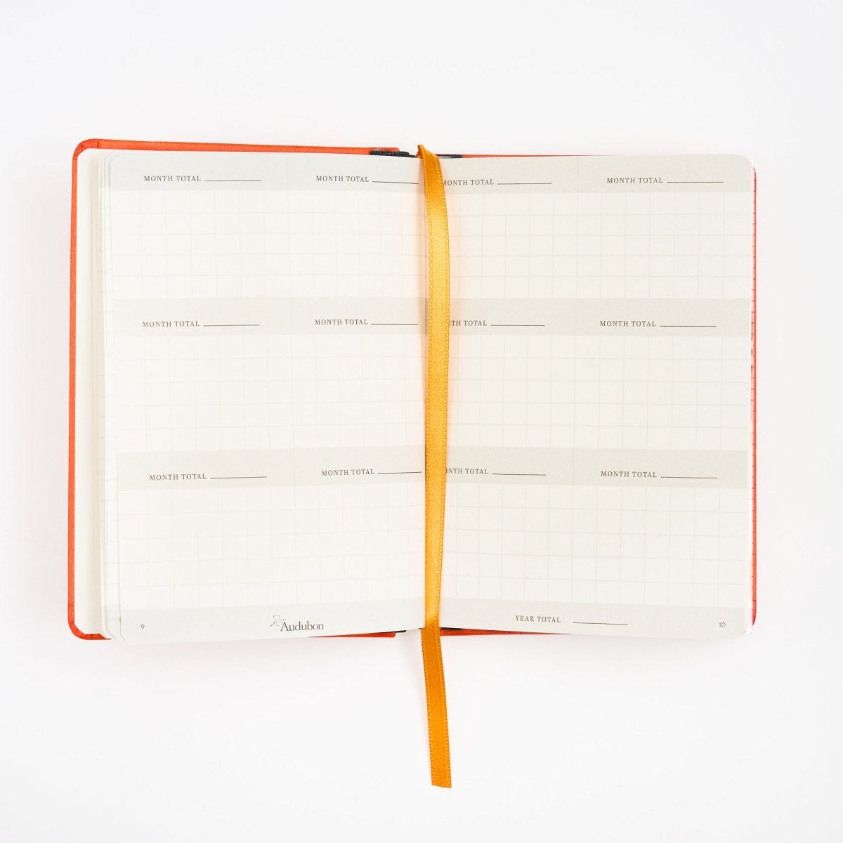 An open monthly birding calendar tracker with white grid pages, an orange cover, and a gold ribbon bookmark, photographed on a clean white background.