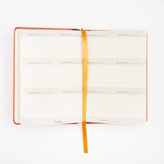 An open monthly birding calendar tracker with white grid pages, an orange cover, and a gold ribbon bookmark, photographed on a clean white background.