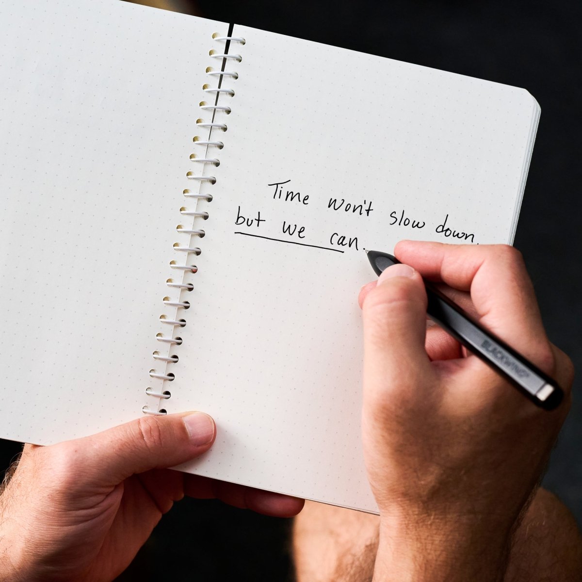 A close-up of a person holding an open spiral notebook with handwritten text 'Time won't slow down, but we can.' on the page and writing with a blackwing pen. The background is dark and out of focus.