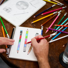 A close-up of a hand drawing colored pencils on the cover of a spiral sketchbook on a wooden table, surrounded by various colored pencils and drawing supplies.