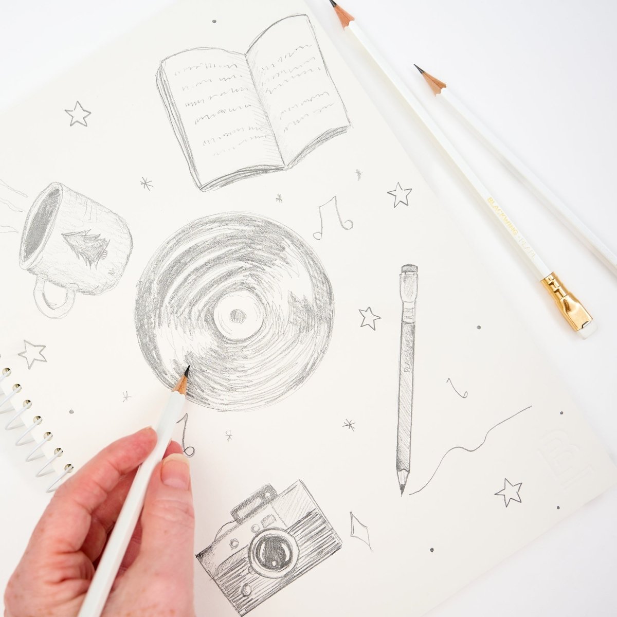 A hand holding a white pencil is sketching on a white spiral-bound notebook. The cover features hand-drawn pencil illustrations of an open book, a mug, a vinyl record, a pencil, a camera, stars, and music notes.