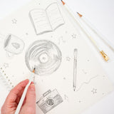 A hand holding a white pencil is sketching on a white spiral-bound notebook. The cover features hand-drawn pencil illustrations of an open book, a mug, a vinyl record, a pencil, a camera, stars, and music notes.