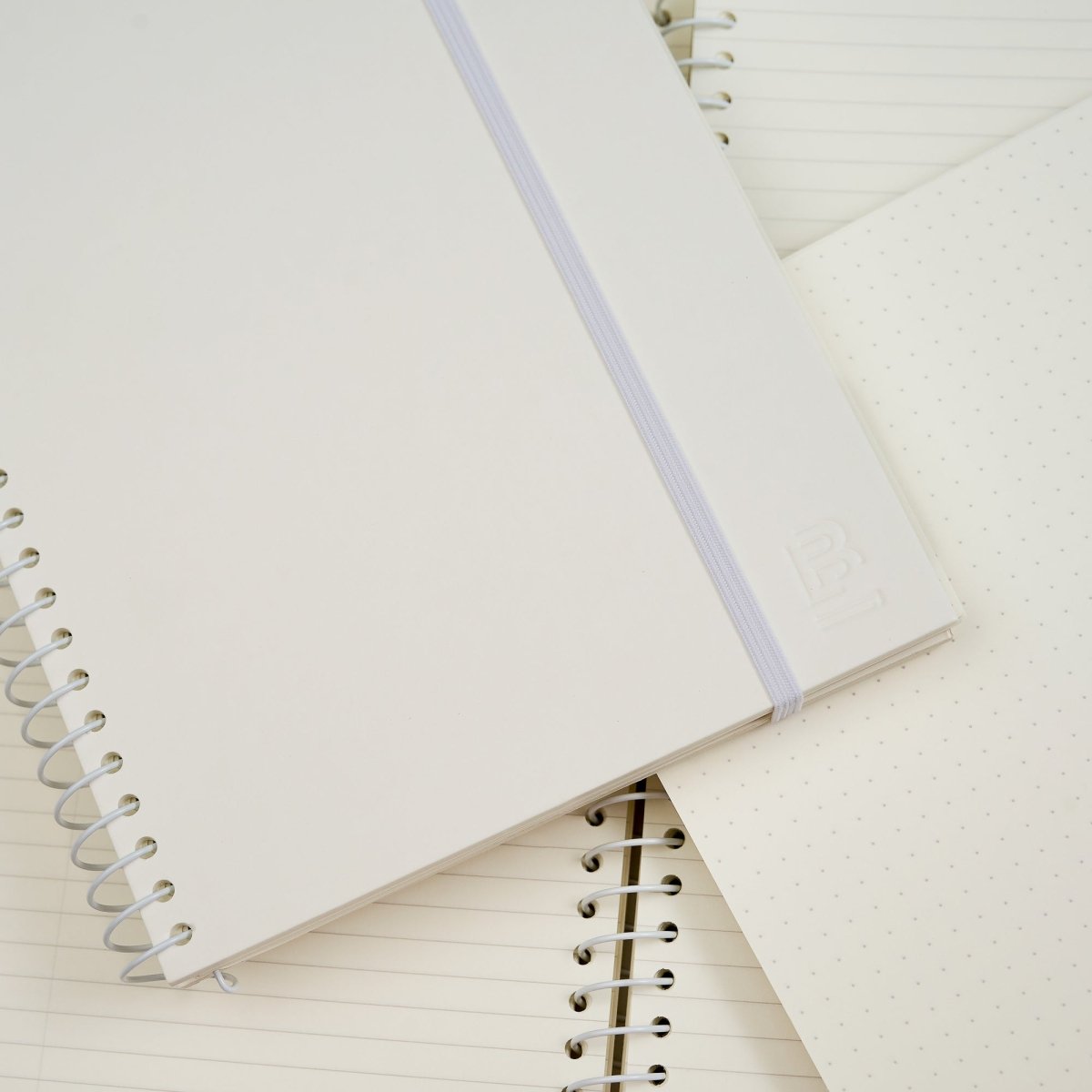 A white spiral-bound notebook with a white elastic band lies diagonally across other notebooks with lined and dotted pages on a clean white background.