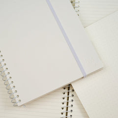 A white spiral-bound notebook with a white elastic band lies diagonally across other notebooks with lined and dotted pages on a clean white background.