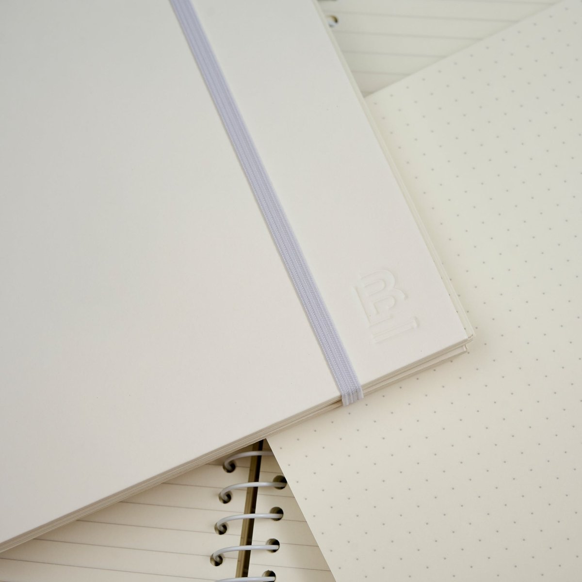 A close-up of a white hardcover notebook with a white elastic closure band and an embossed 'B' logo on the cover, layered over other notebooks with dotted and lined pages.