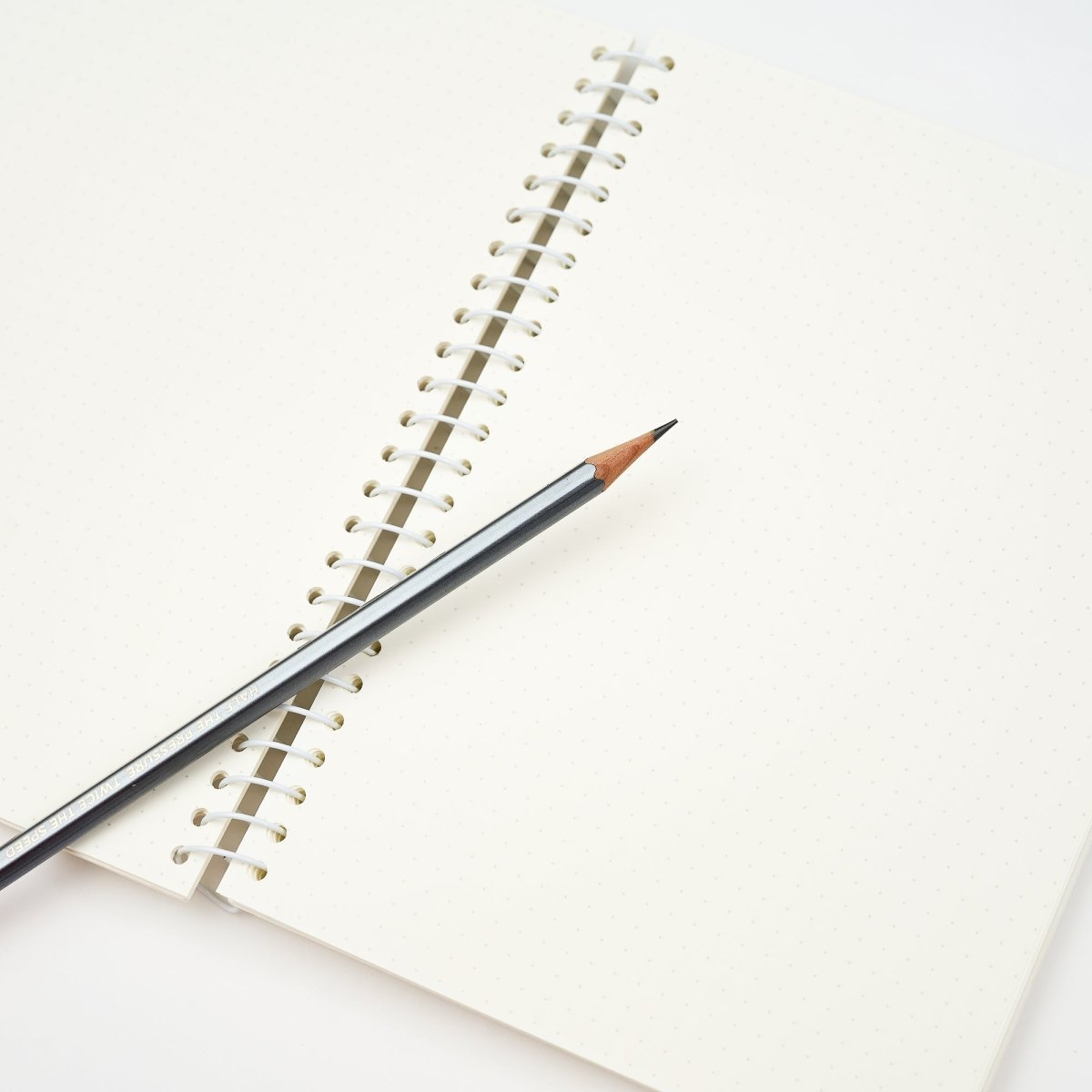 A close-up of an open spiral-bound notebook with blank white pages, with a dark gray pencil resting diagonally across the spiral binding.