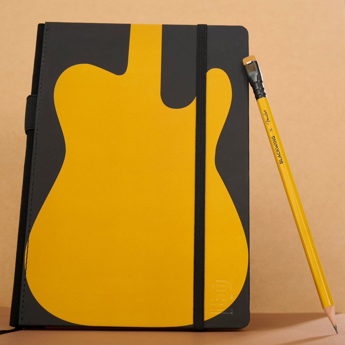 A notebook with a black cover featuring a large yellow guitar-shaped graphic to symbolize the Fender Butterscotch guitar, shown standing upright next to a black and yellow pencil on a beige background.