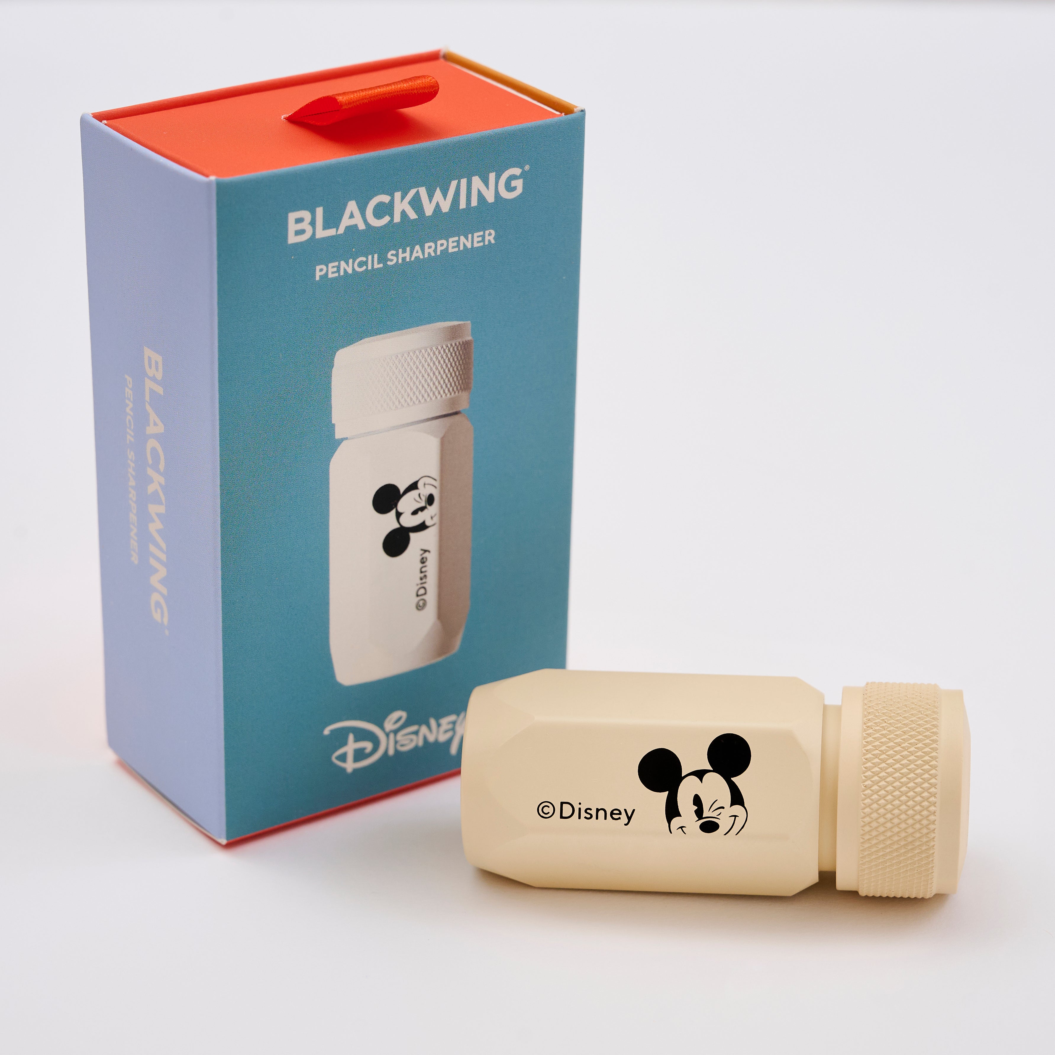 A Disney Blackwing pencil sharpener in a cream color with a black Mickey Mouse face logo, shown next to its blue and red packaging box.