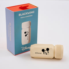 A Disney Blackwing pencil sharpener in a cream color with a black Mickey Mouse face logo, shown next to its blue and red packaging box.