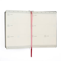 An open weekly Blackwing planner with blank day slots on the inside pages, held open by a pink ribbon bookmark, shown on a white background.