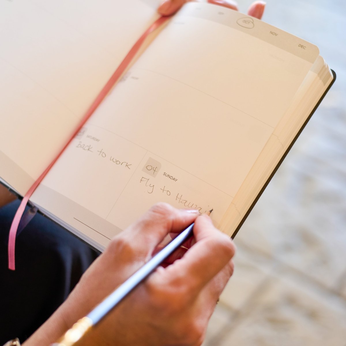 A close-up of a person writing in a Blackwing planner  with a Blsckwing 602 pencil, held open by another hand. The notebook has a cream page layout with a pink ribbon bookmark and a weekly planner section visible.