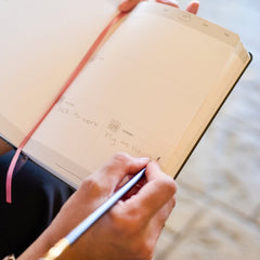 A close-up of a person writing in a Blackwing planner  with a Blsckwing 602 pencil, held open by another hand. The notebook has a cream page layout with a pink ribbon bookmark and a weekly planner section visible.