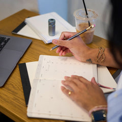 A close-up lifestyle shot of a person writing in a Blackwing planner with a pencil, set on a wooden desk alongside a laptop, notebook, and an iced coffee.