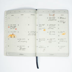 Open Blackwing Planner showing a filled-in monthly calendar spread with handwritten notes, small drawings, and a grey ribbon bookmark against a white background.