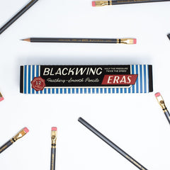 A flat-lay of a Blackwing Eras graphite smooth pencil box on a white background, surrounded by several black pencils with gold ferrules and pink erasers.