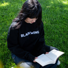 Woman sitting in the grass reading a book, wearing a black crewneck sweatshirt with 'BLACKWING' embroidered in white across the chest.
