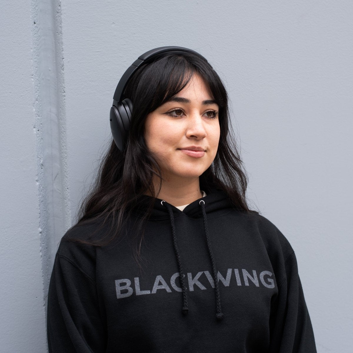 Person wearing black headphones and a black hoodie with 'BLACKWING' logo against a gray wall.