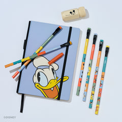 A Disney-themed stationery set on a light gray background, featuring a light blue notebook with Donald Duck artwork, six colorful pencils, a beige Mickey Mouse sharpener, and a red pencil point guard.