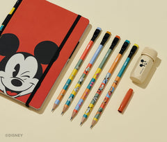 A Disney stationery set arranged on a warm cream background, including a red notebook with a Mickey Mouse face, five colorful pencils, a red pencil guard, and a beige sharpener with a Mickey Mouse head print.