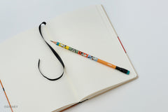 An open blank notebook with a black ribbon bookmark and a decorative colorblock pencil featuring colorful Goofy illustrations, photographed on a clean white background.